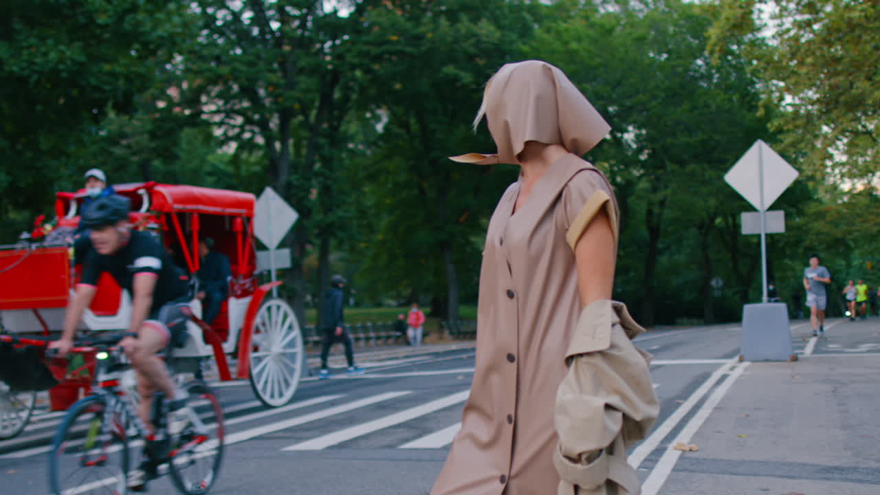 New York, USA, 25 maggio 2025: Donna in abito beige con carrozza trainata da cavalli a New York. Scena di strada di una donna che indossa un cappotto senza maniche e un foulard che passa accanto a una carrozza trainata da cavalli in un parco.