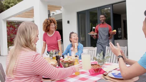 African-american-man-serving-food-to-diverse-group-of-friends-at-dinner-table-in-garden,-slow-motion