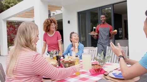 Hombre-Afroamericano-Sirviendo-Comida-A-Un-Grupo-Diverso-De-Amigos-En-La-Mesa-Del-Jardín,-Cámara-Lenta