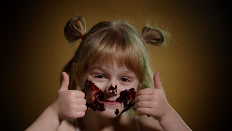 Portrait-of-teen-smiling-child-kid-girl-smears-face-with-melted-chocolate-and-showing-thumbs-up-sign