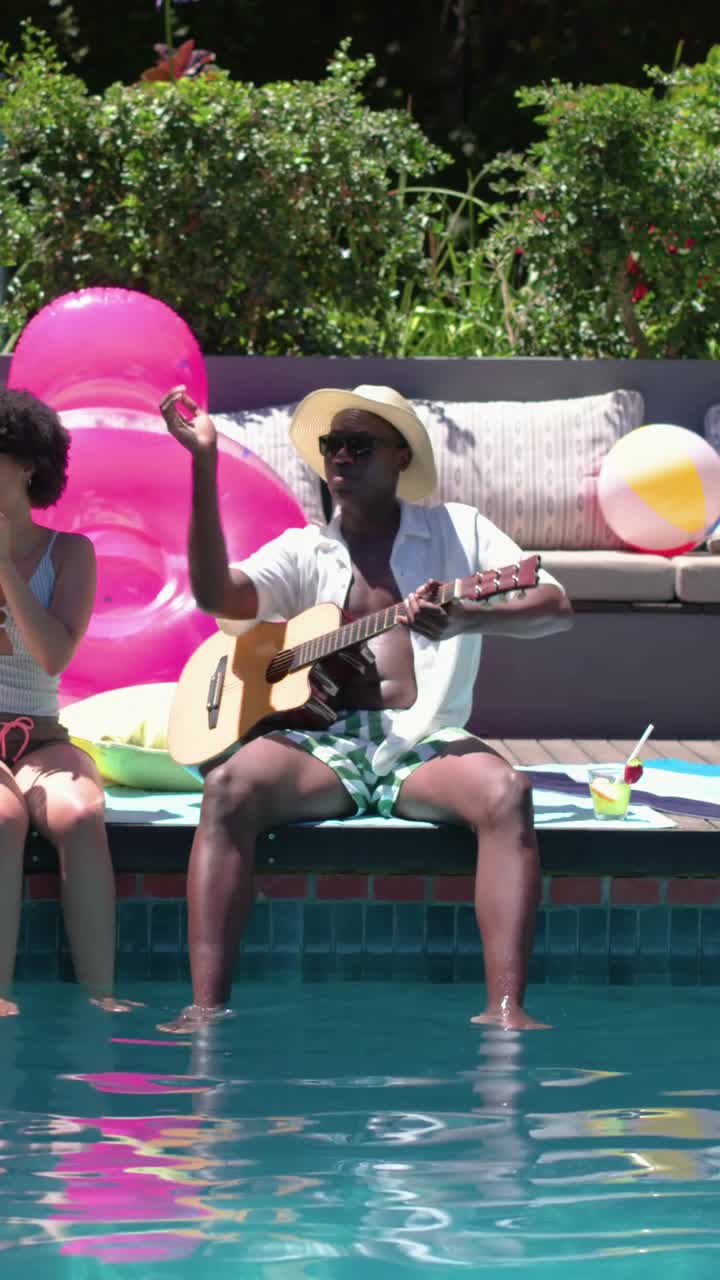Video verticale: Un uomo con cappello di paglia che saluta mentre suona la chitarra acustica a bordo piscina per un amico che applaude.