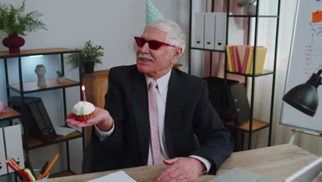 senior businessman celebrating lonely birthday in office, blowing candle on small cake making a wish