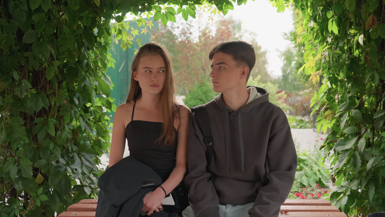 Due adolescenti seduti su una panchina in un'alcova verdeggiante, lei con un abito nero e lui con una felpa grigia, si scambiano sguardi dolci e un silenzio misurato. Un arco ricoperto di viti incornicia un giardino di periferia, la luce estiva crea un controluce