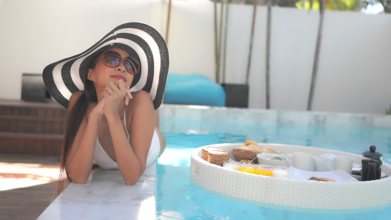 Luxury Asian woman lying at the edge of swimming pool resort next to the floating tray with breakfast In Thailand