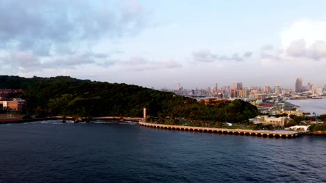 A-lush-hillside-by-the-sea-with-city-skyline-at-sunset,-aerial-view