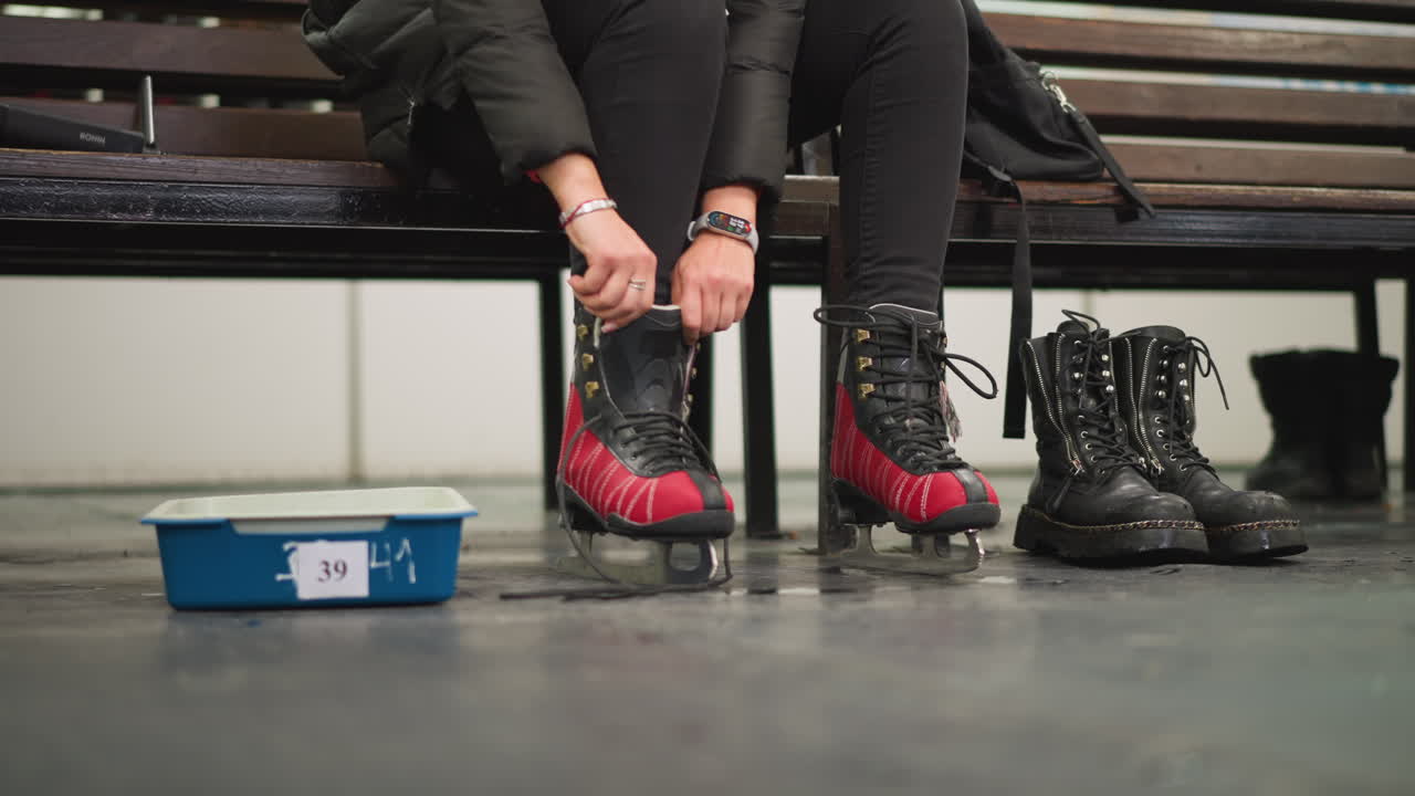 Lady sitting on bench putting on red black ice skates, wearing black pants and dotted white socks, with combat boots on floor, preparing for skating session indoors with focus on skates moment