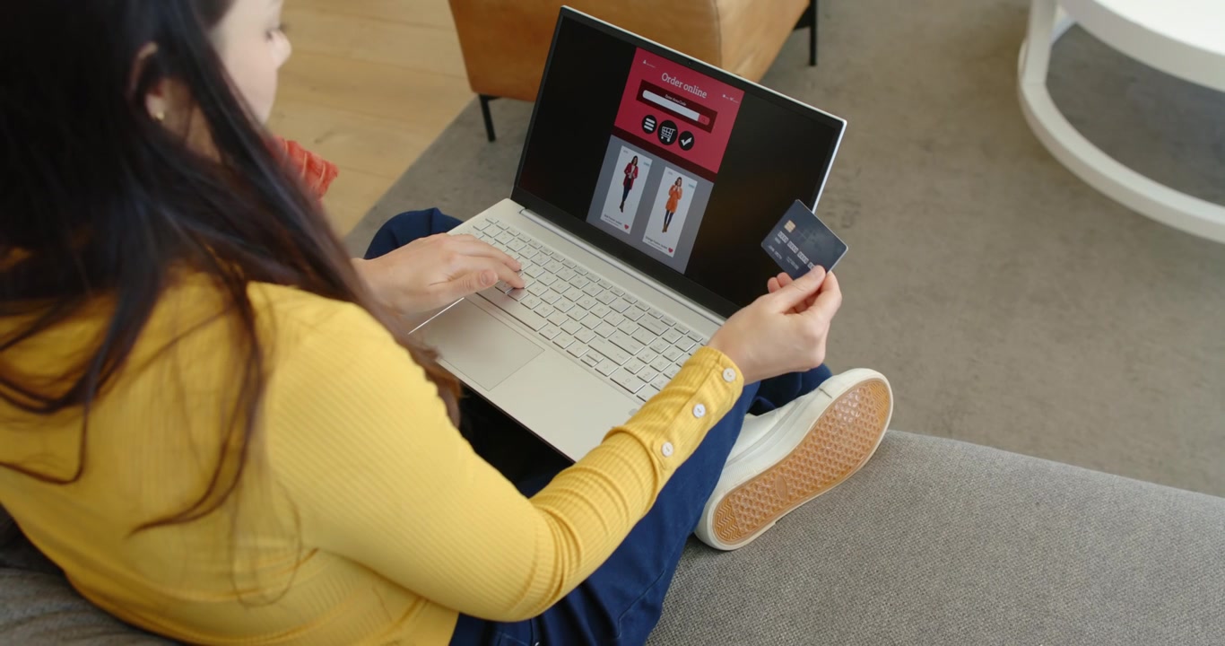 Mujer adulta asiática sentada en un sofá gris, usando una laptop para mostrar opciones de vestidos y sacando una tarjeta de crédito.
