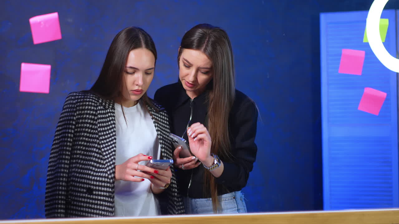 Two brunettes with long straight hair are looking at phone. Bloggers check the recorded video content.