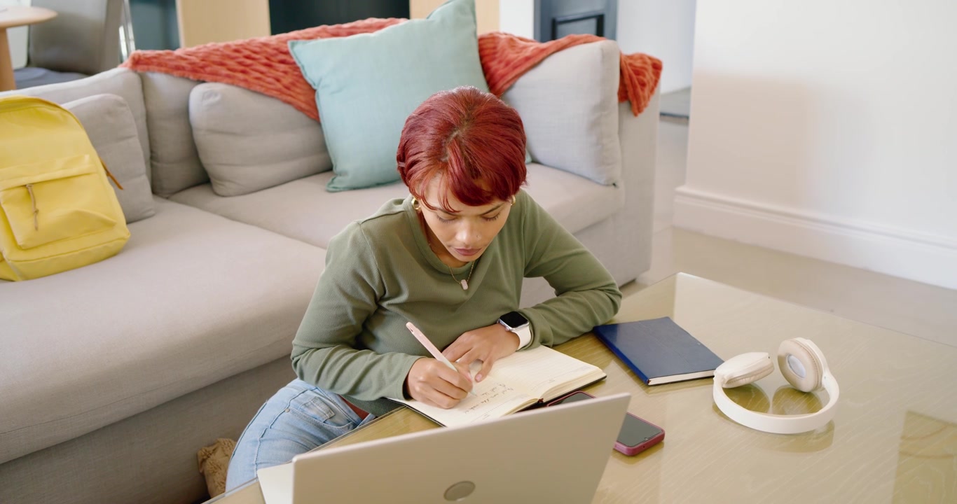 African American woman glancing at laptop, studying on floor at table, writing in notebook with pen