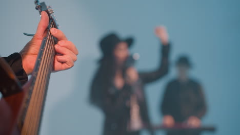 close view of a faceless guitarist playing a guitar against a white background with a blurred singer and keyboardist in the background. the guitarist's hand is focused as they strum the strings