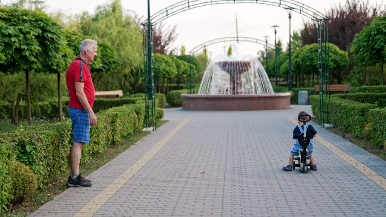 Grandpa shows his grandson how to ride a scooter in the park. Grandfather pretends to run away so the baby could follow him.