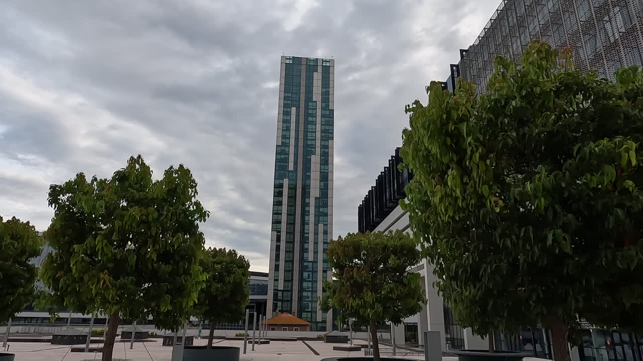Timelapse drammatico di un moderno grattacielo di vetro che si erge al di sopra di alberi verdi e lussureggianti a Roma, Italia. L'architettura urbana moderna contrasta con la natura sotto un cielo nuvoloso dinamico