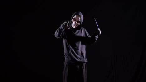 hombre aterrador en la sudadera con capucha con máscara de halloween haciendo un gesto de despedida mientras sostiene un cuchillo en el fondo negro