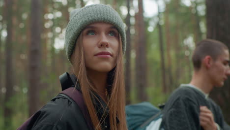 girl wearing beanie and jacket looks around thoughtfully in forest with soft expression while standing beside companion carrying sleeping bag, surrounded by tall trees and green