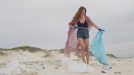 mujer caucásica con guantes de látex recogiendo basura de la playa usando un palo de agarre
