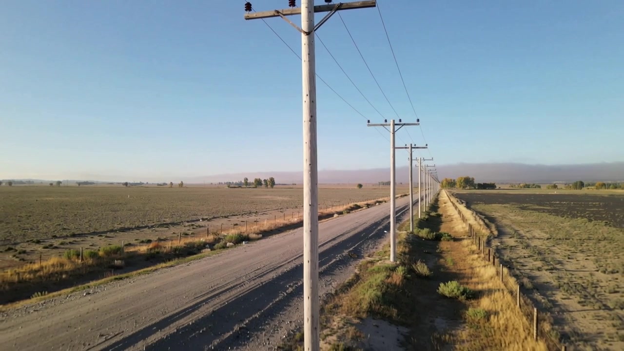 Cámara comenzando a avanzar por un camino sin pavimentar, revelando postes de servicios, una valla y colinas distantes.