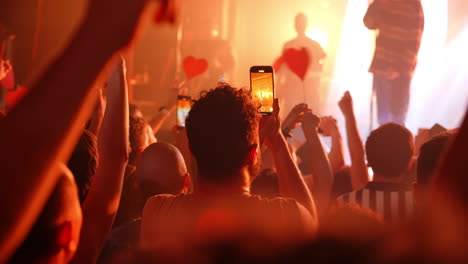enthusiastic concertgoers holding phones and heart shaped signs, immersed in vibrant live music performance with dynamic lighting, capturing electric moment of collective joy and unity