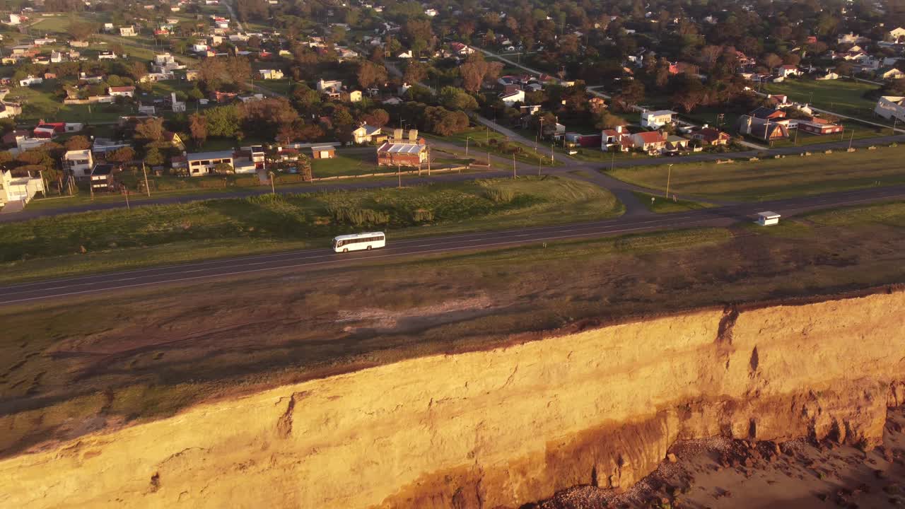 일몰 동안 절벽과 바다 옆 해안 도로를 주행하는 흰색 버스의 공중 추적 샷
