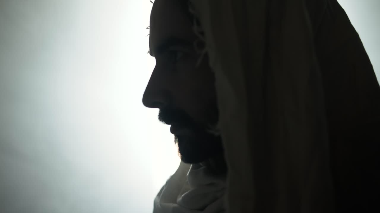 Closeup of the face of Jesus Christ resurrected on Easter Sunday inside empty tomb in bright, shining light, wearing white hood and robe. Cinematic Christian visual.
