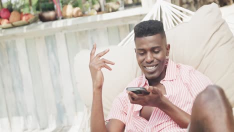 Fröhlicher-Afroamerikanischer-Mann,-Der-In-Der-Hängematte-Sitzt-Und-Am-Sonnigen-Strand-Auf-Dem-Smartphone-Spricht,-Zeitlupe