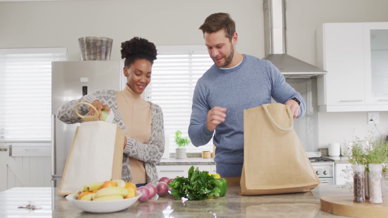 video de una feliz pareja diversa desempaquetando comestibles en la cocina