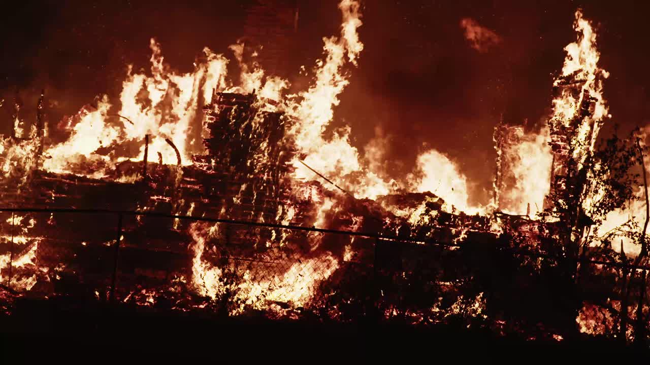 Intense flames rip through a large structure behind a fence as the night sky glows orange with smoke and fire