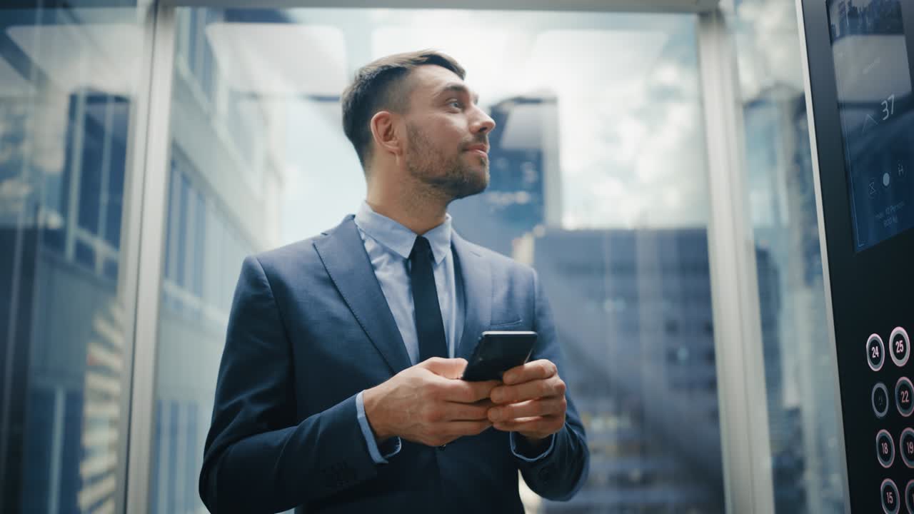 hombre de negocios exitoso montando un ascensor de vidrio a la oficina en un centro de negocios moderno. hombre feliz y guapo sonríe mientras usa el teléfono inteligente, escribe mensajes de texto, revisa las redes sociales y los correos electrónicos de trabajo en un ascensor.