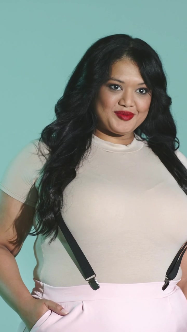 Vertical video: Camera cueing model posing, turning head and smiling on teal set with suspenders