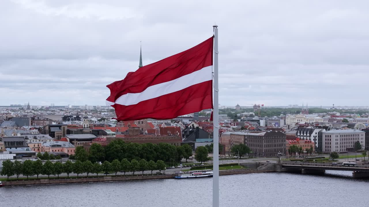Massive Latvian flag flutters on cloudy day above Riga with Old Town panorama