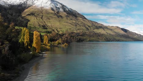 slowmo - flying over beautiful blue lake wakatipu, queenstown, new zealand with mountains fresh snow, clouds, blue sky autumn trees in the background - aerial drone