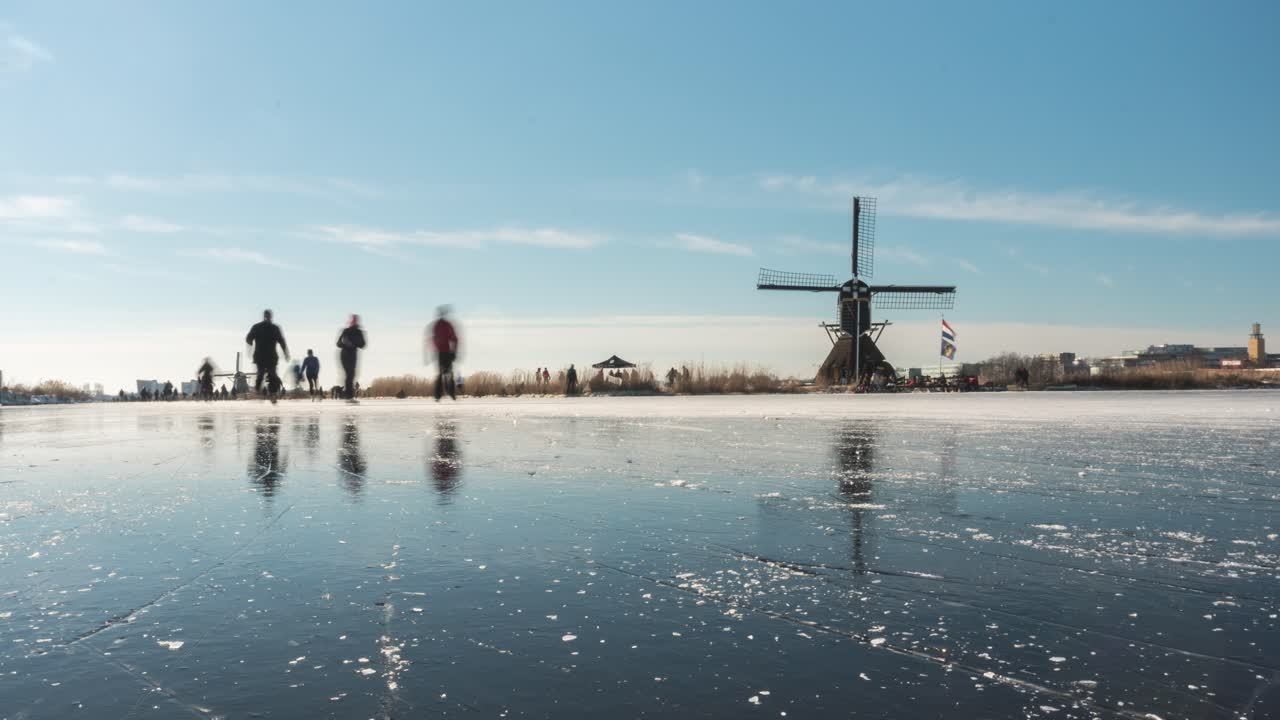 Time lapse of Dutch ice skaters on frozen canal beside old windmill, slider shot