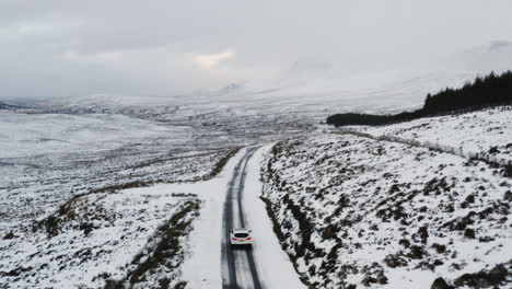 stunning aerial overview of car driving along barren single lane road covered with snow on forest edge