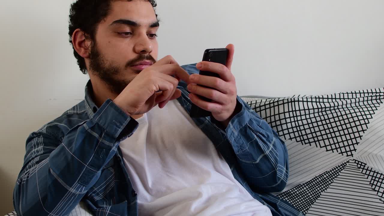 Young man searches for content on his mobile, rests on the sofa in the living room of his home.