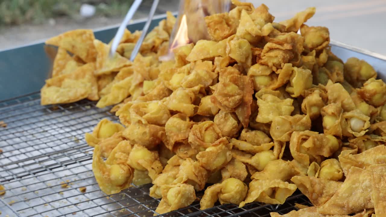 Hand selects crispy fried wontons from tray at outdoor Thai street food market, natural daylight