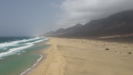 toma aérea de grandes olas rompiendo en la larga playa blanca de cofete con hermosas montañas en el fondo en fuerteventura, isla canaria