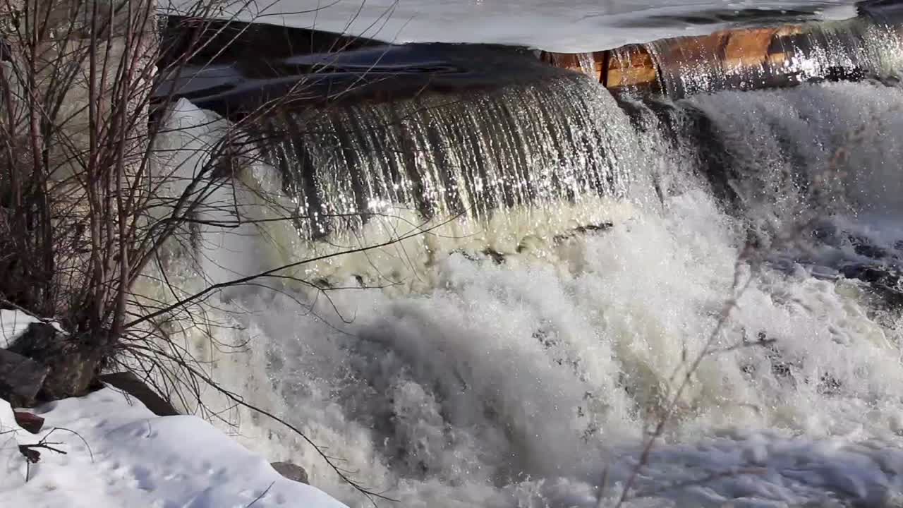 cold rushing water pours over frozen dam. Stationary close up