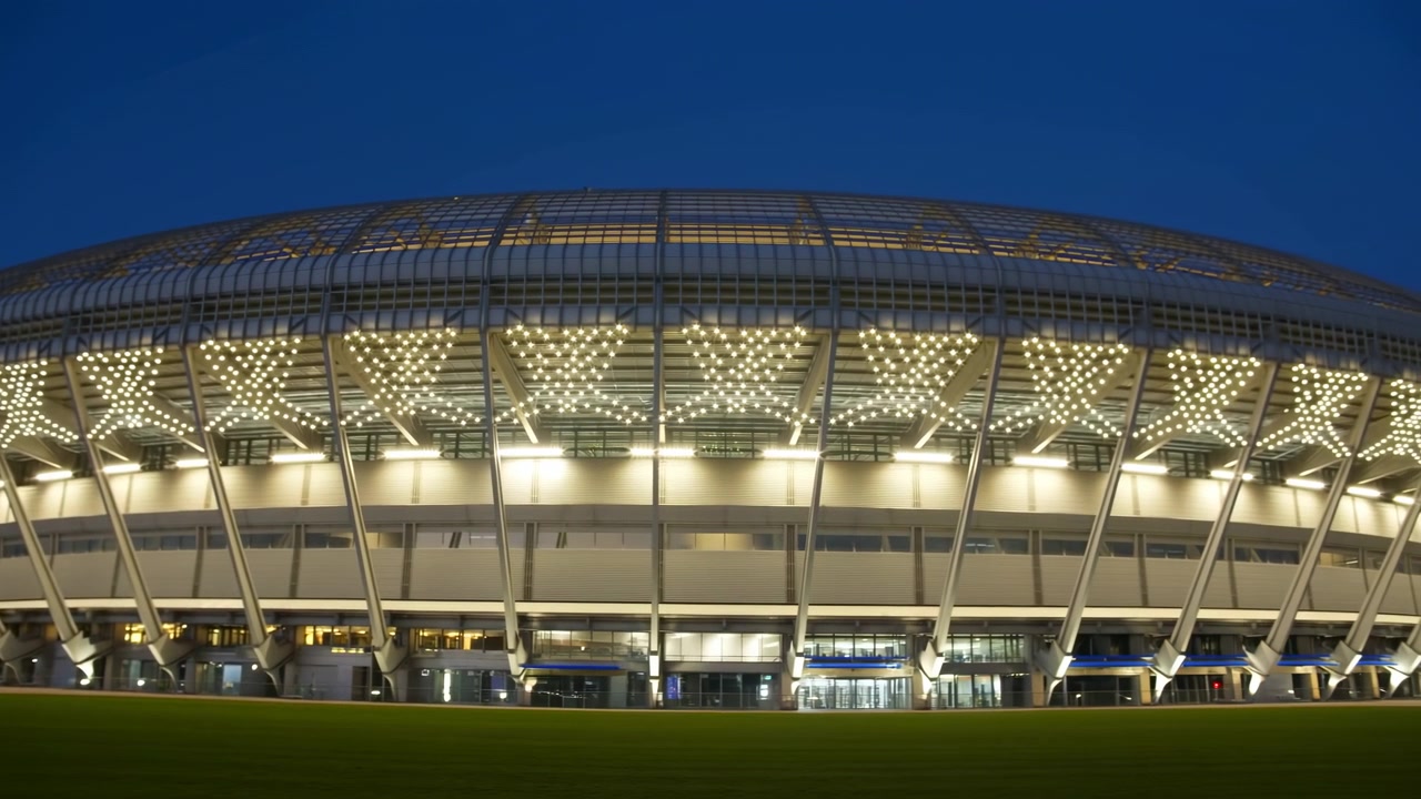 Beleuchtete moderne Stadionaußenansicht bei Nacht, die eine geschwungene beleuchtete Fassade und ein strukturelles Gitterwerk zeigt, leuchtend.