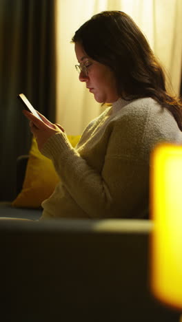 vertical video of young woman spending evening at home sitting on sofa with mobile phone scrolling through internet or social media