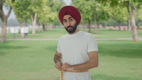 happy sikh indian man measuring waist using inch tape in park