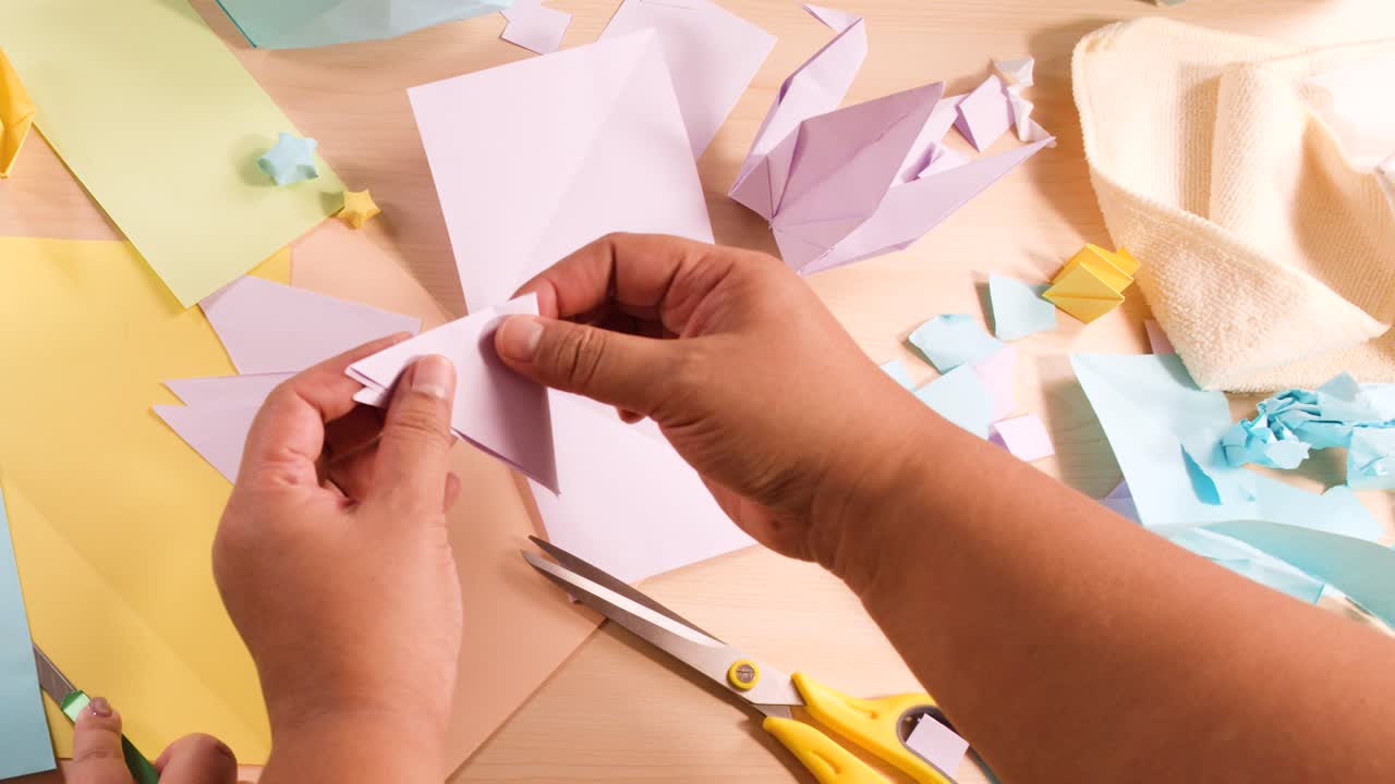 Primo piano dall'alto di mani che piegano carta viola creando forme origami su una scrivania piena di oggetti