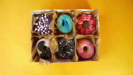 two hands present a box containing six assorted donuts, each uniquely decorated with toppings like marshmallows, sprinkles, cookies, and candy, set against a bright yellow background.