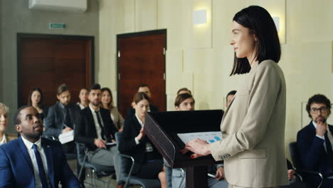 rear view of businesswoman speaking on the podium in a conference room and demonstrating video presentation on the screen behind her