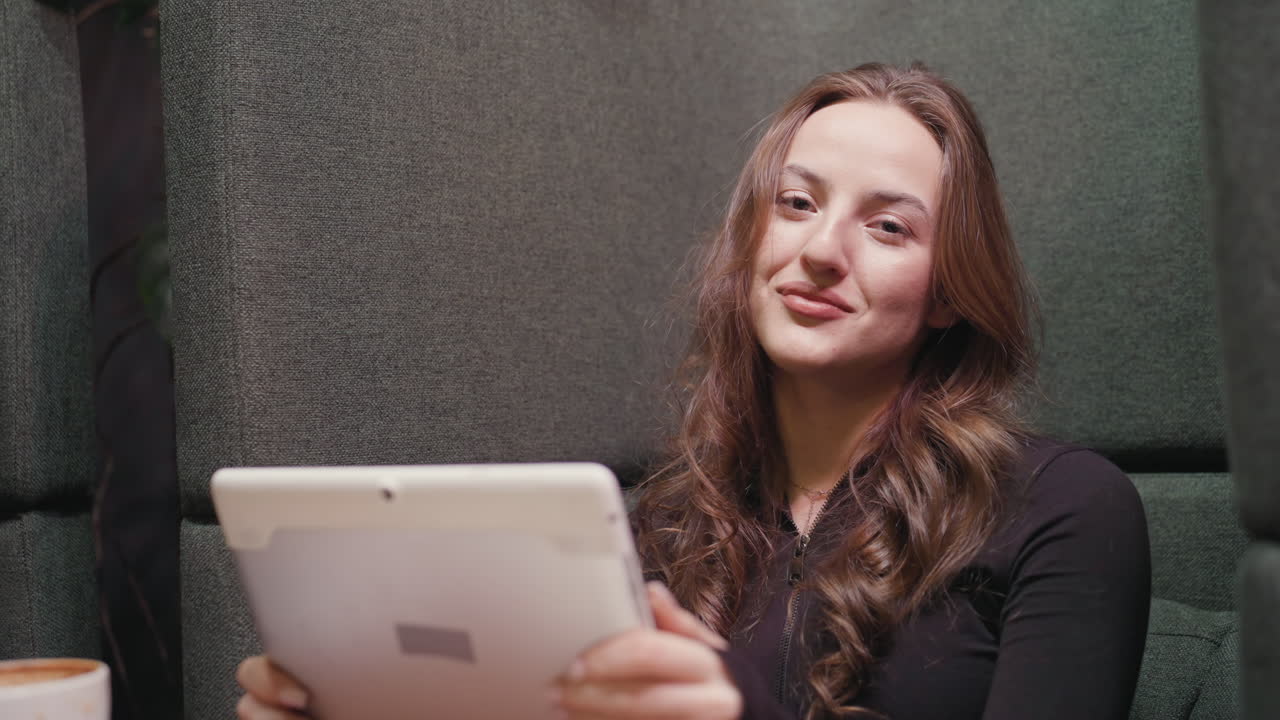 Close view of relaxed lady with long wavy hair holding digital notepad and looking at camera with soft expression, seated against dark cushioned wall in indoor environment with latte in foreground