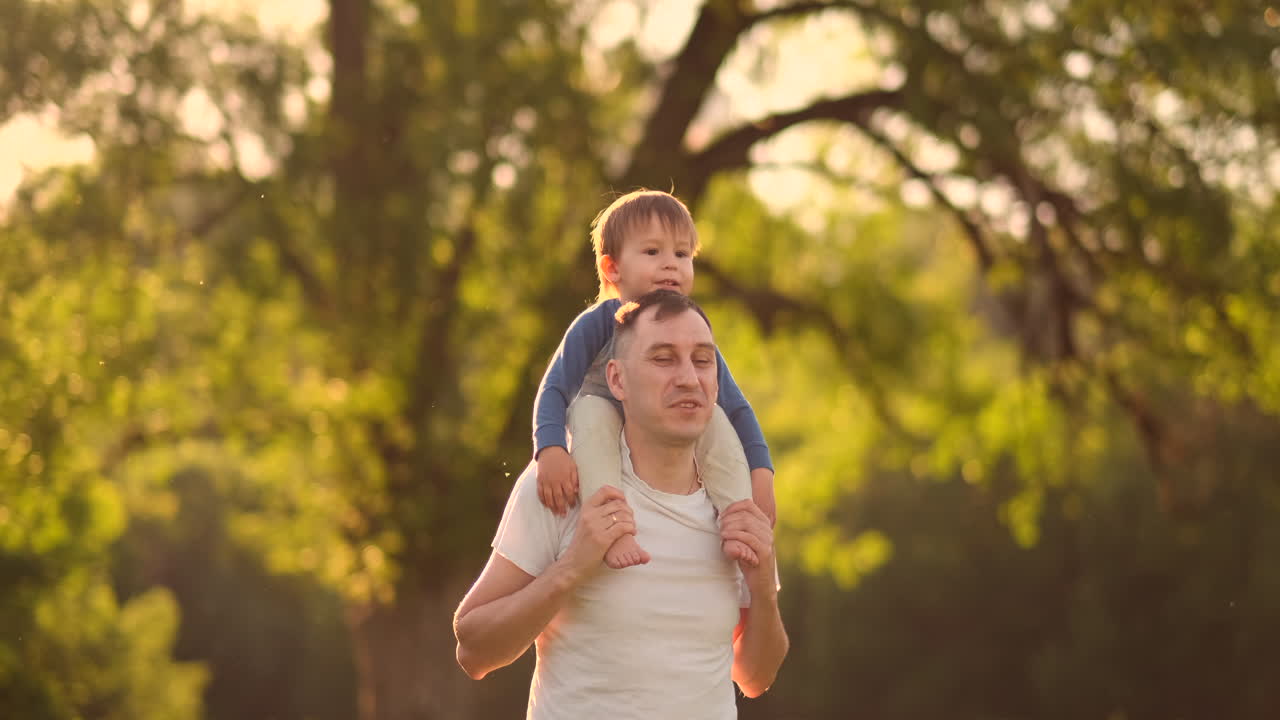 padre amoroso sonríe caminando con el niño sentado en el cuello al atardecer en un prado en verano en cámara lenta.