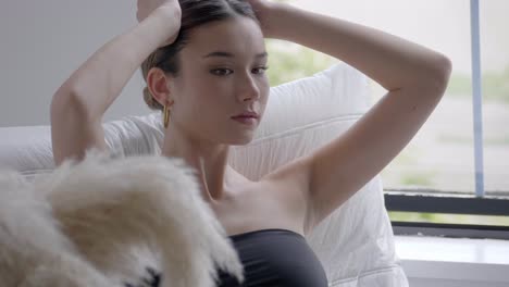 Beautiful-Young-Woman-In-Black-Tube-Top-Sitting-On-A-White-Couch-Holding-Her-Head-With-Both-Hands-Relaxing-Inside-A-Hotel-Room