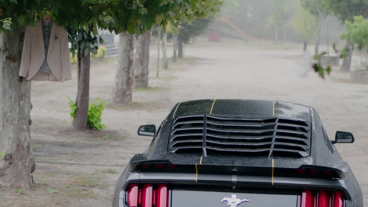 Black sports car parked under a tree with a beige jacket hanging from a branch nearby