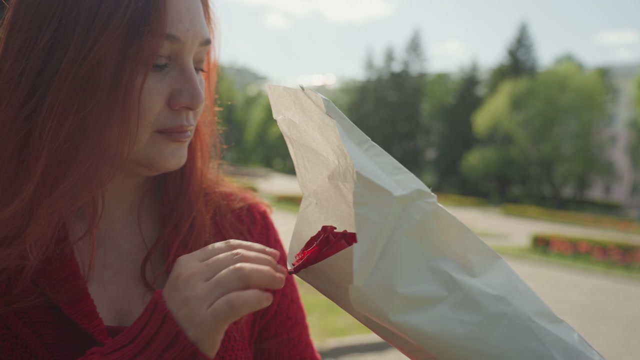 Primo piano di una donna con lunghi capelli rossi che indossa un maglione di lana, mentre apre con cura un foglio bianco per rivelare dei fiori. La scena è ambientata all'aperto in una giornata di sole, con alberi verdi e in lontananza.