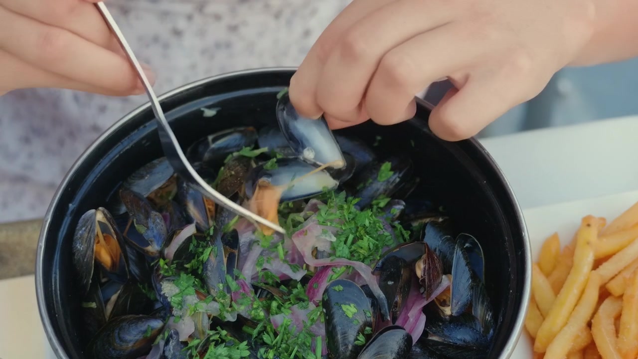 Close-up of hands stirring a pot of mussels with a fork, a plate of fries beside it, Cote d'Azur, France