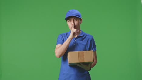 mensajero masculino asiático en uniforme azul mostrando un gesto de silencio y sacudiendo la cabeza mientras entrega un cartón en el fondo de la pantalla verde en el estudio