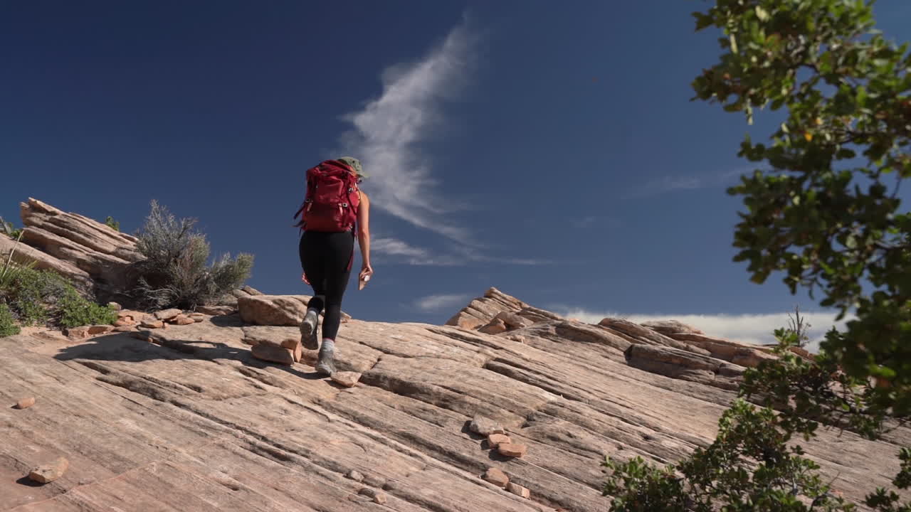 espalda de mujer con mochila caminando sobre una colina rocosa en el desierto bajo el cielo azul, cámara lenta